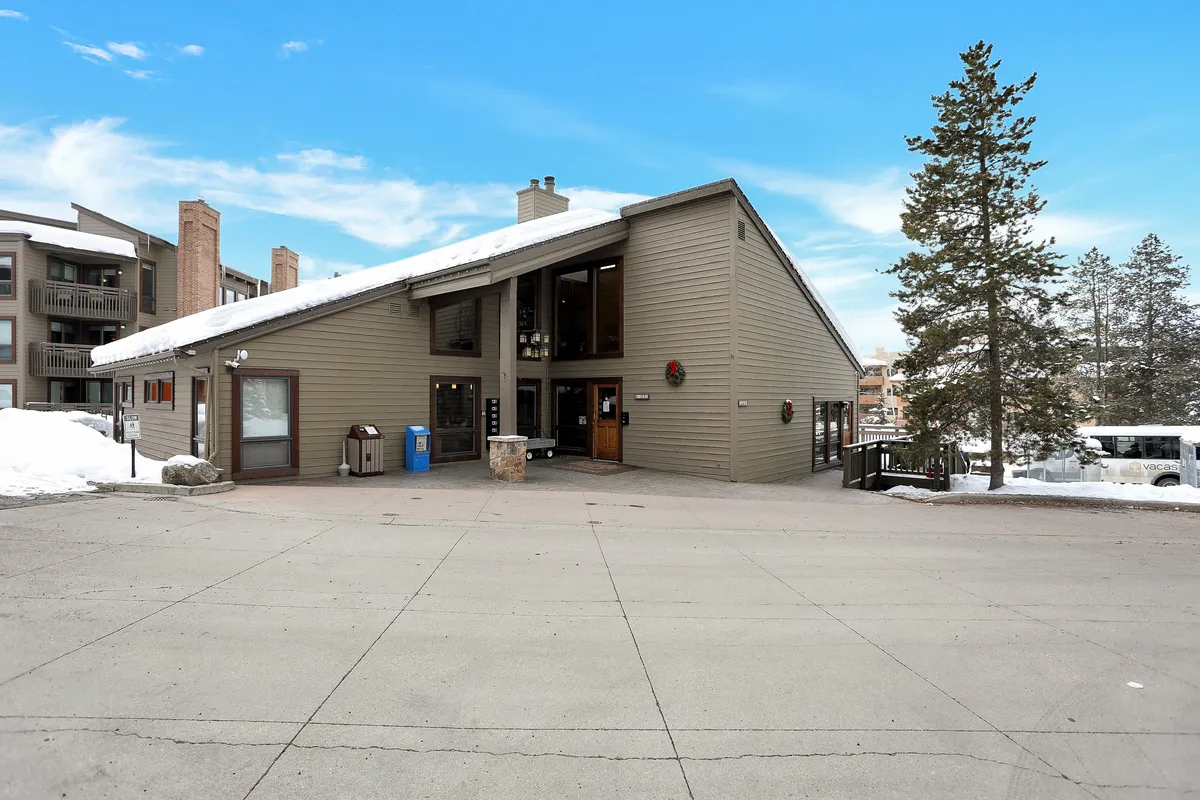 The image shows the building entrance surrounded by snow-covered ground and trees, with a visible parking area. The snowy landscape and welcoming entryway evoke the convenience of returning home after a day on the slopes and the proximity to ski activities.