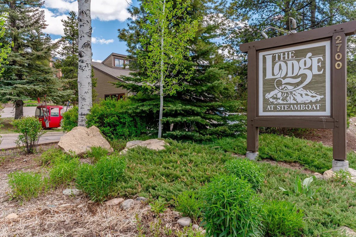 The property’s exterior features a lodge sign, landscaped grounds, surrounding trees, and a red vehicle, reinforcing easy access to the neighborhood. The welcoming entryway and communal vibe hint at walkable convenience to area amenities.