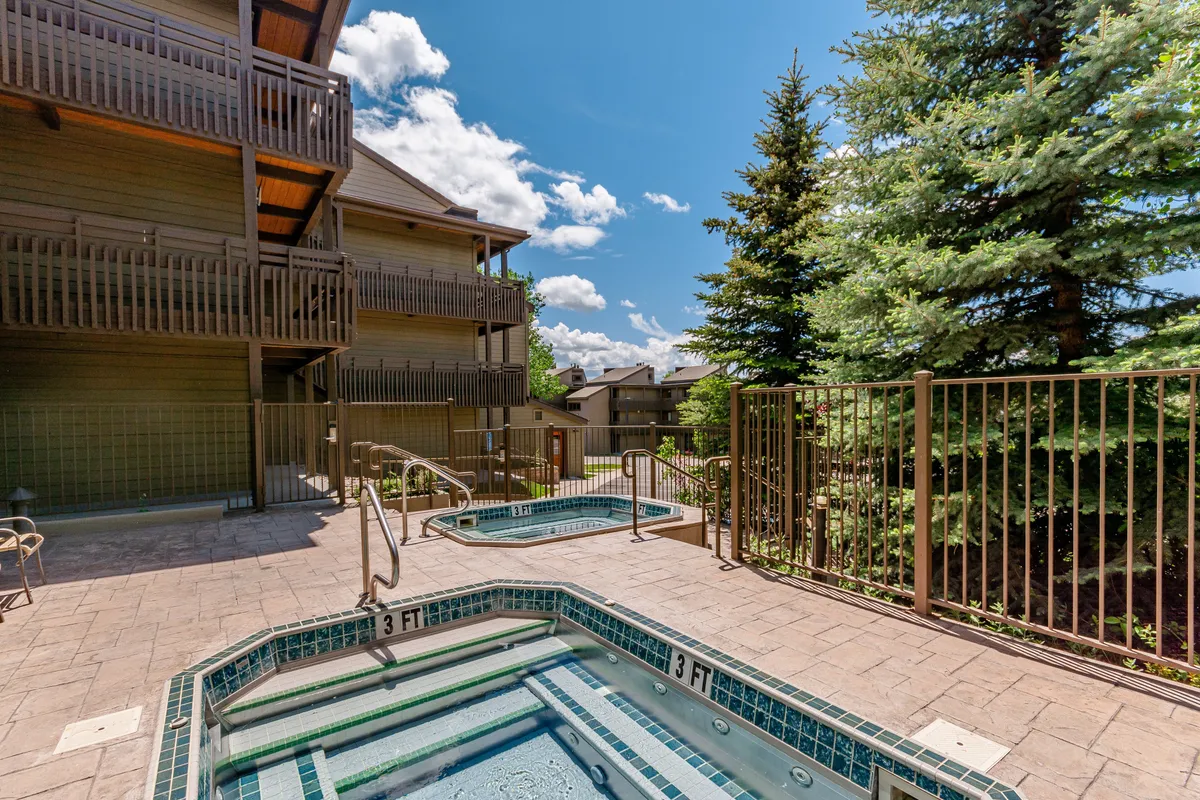 Two hot tubs nestled within a landscaped outdoor area, framed by greenery and the balconies of nearby buildings. The scene conveys a sense of relaxation and social gathering, with the hot tubs inviting guests to unwind in the fresh mountain air.
