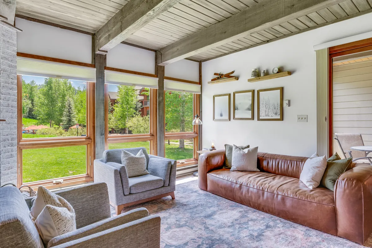 A welcoming living room with large windows that offer a clear view of the outdoors. The space features a brown leather sofa, light grey armchairs, and a decorative wall shelf with framed pictures, creating a cozy environment ideal for watching the changing seasons. The room captures both relaxation and socialization, with strong visual connection to the landscape beyond.
