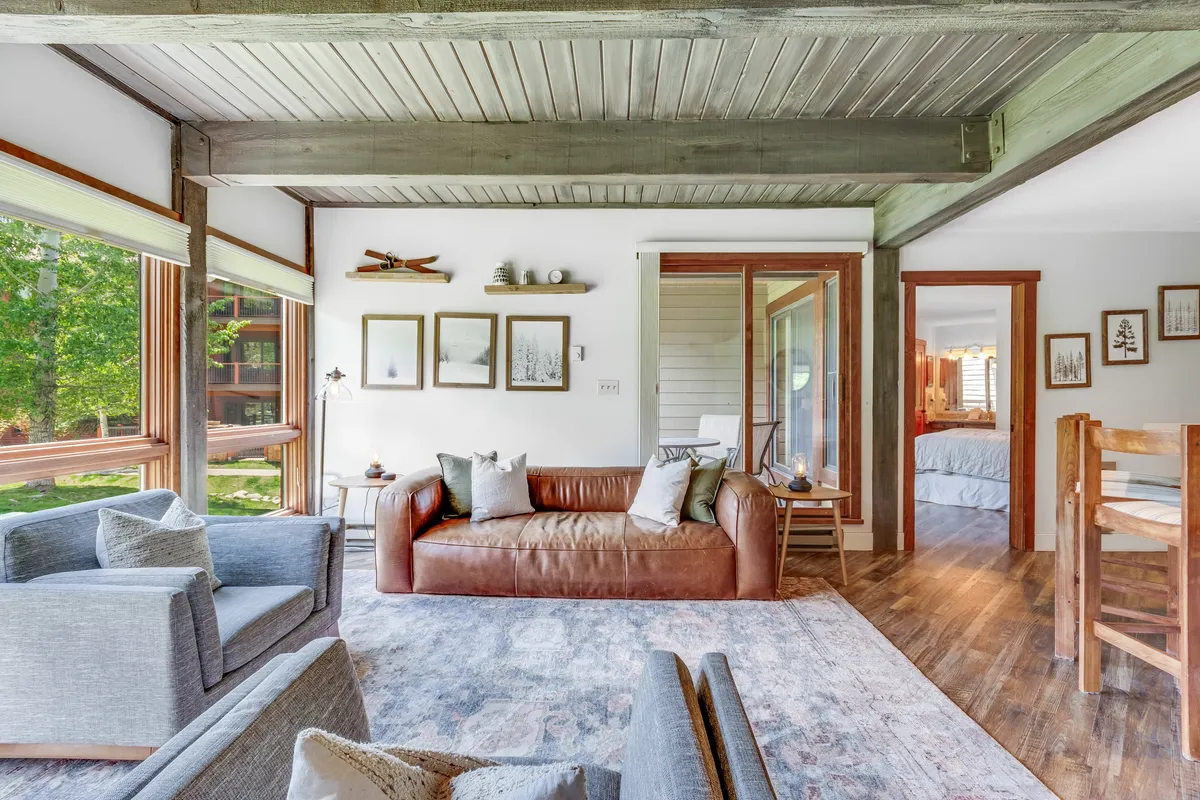 A warm living area featuring a leather sofa positioned near large windows that invite natural light and offer glimpses of the outdoors. The room includes decorative wall art and a wooden dining area, creating a comfortable space perfect for relaxing, socializing, or soaking in nature views.