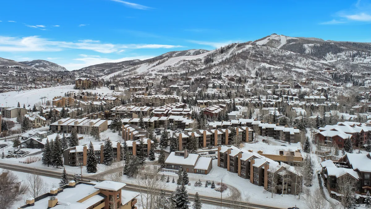 This image captures snow-covered rooftops, distant mountains, tree-lined streets, and the visible ski slopes in the background. The serene winter landscape and the proximity of the slopes illustrate the allure and excitement of a Steamboat ski getaway.