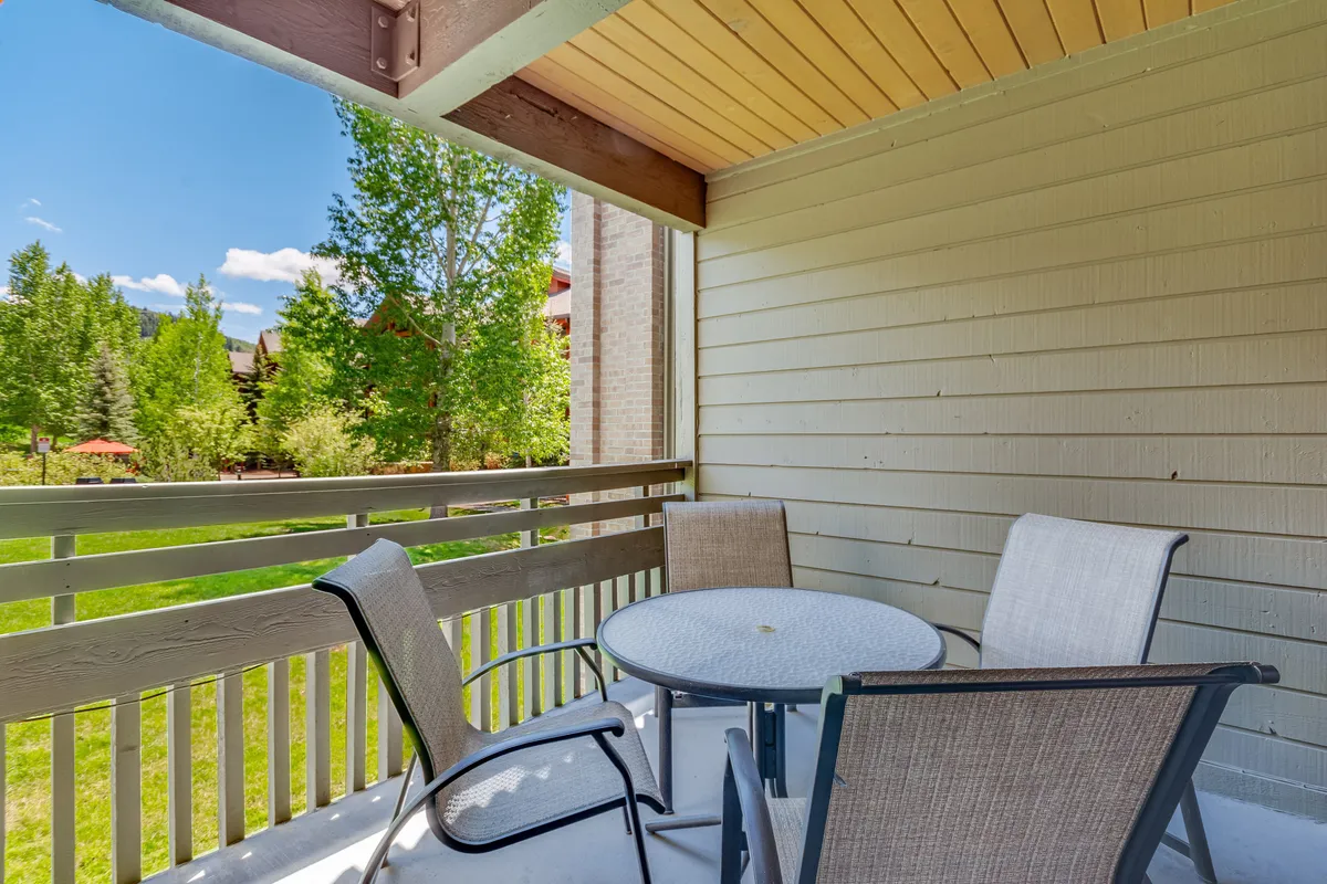 An inviting exterior balcony space with a round table and four chairs, surrounded by lush greenery. The scene suggests a tranquil spot for morning coffee, an al fresco meal, or evening drinks as the sun sets, with the balcony railing framing the natural surroundings.