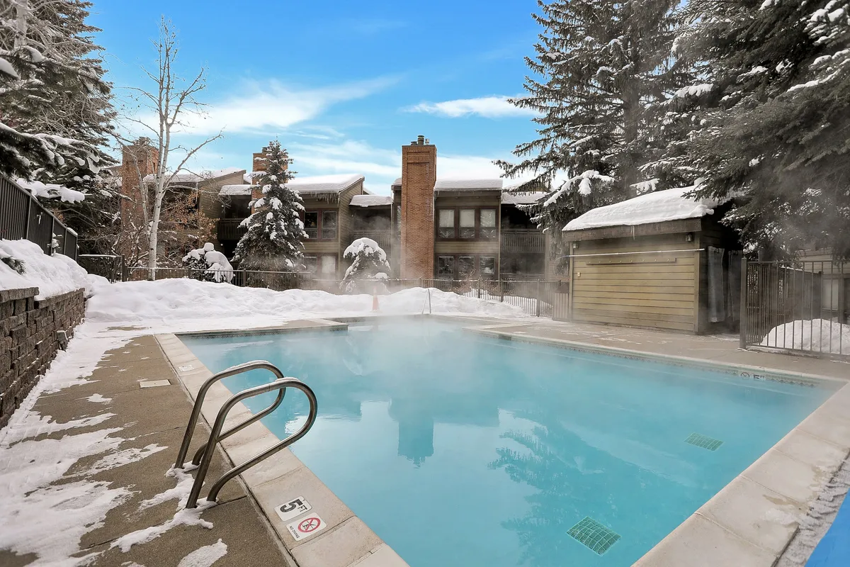 A swimming pool surrounded by snow, with steam rising into the winter air and trees framing the scene. The contrast of the warm pool against the snowy landscape highlights unique après-ski relaxation options on the property.