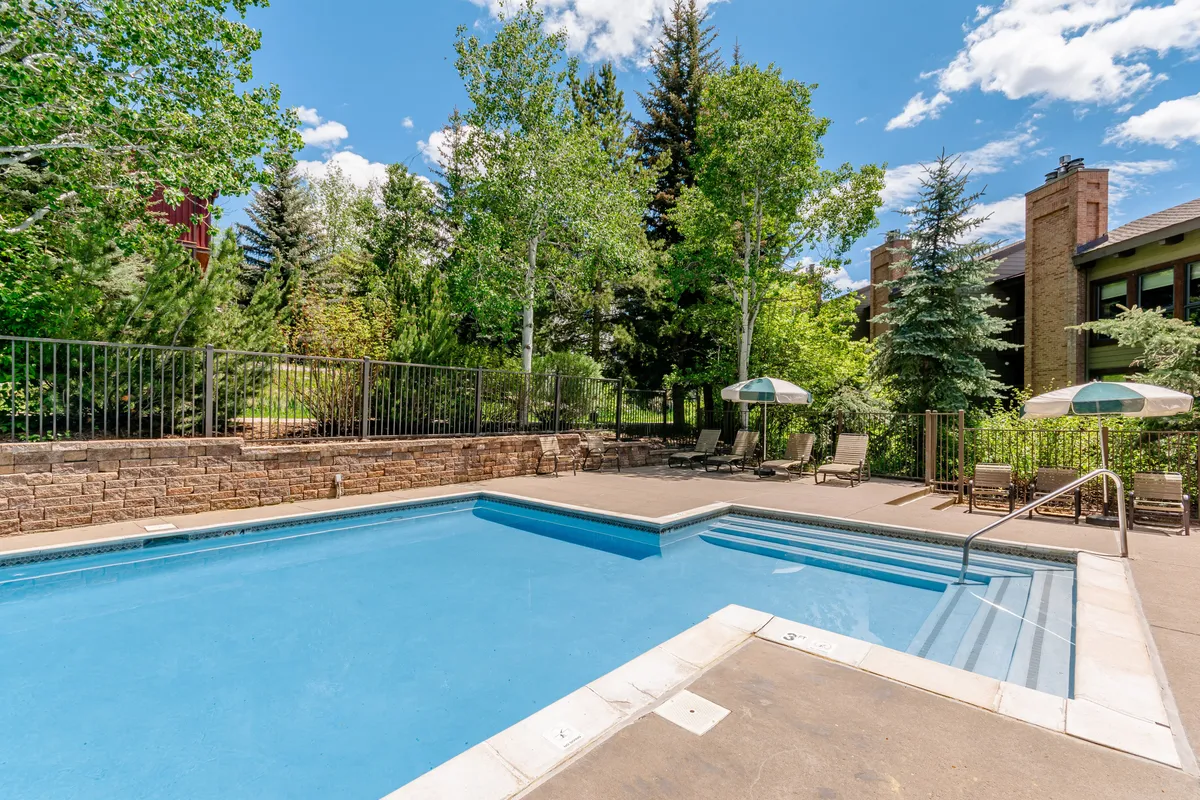 A sparkling outdoor swimming pool surrounded by lush trees and several lounge chairs arranged for relaxation. The scene suggests a tranquil environment ideal for swimming, sunbathing, or enjoying a quiet moment poolside. The pool area is bright and open, providing a resort-style atmosphere.