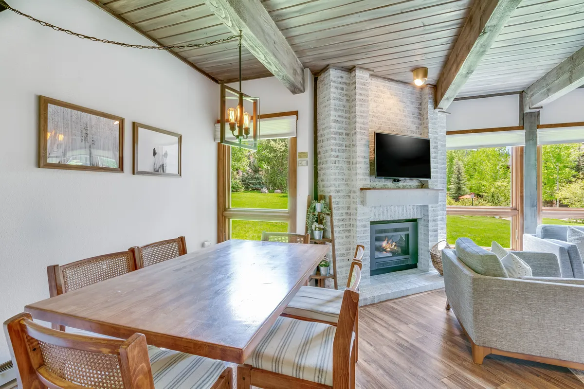 A bright, inviting open-concept living/dining space featuring a central fireplace, a dining table set for gatherings, a plush sofa, and expansive windows. The setting is ideal for family meals, evenings by the fire, and enjoying the surrounding scenery.